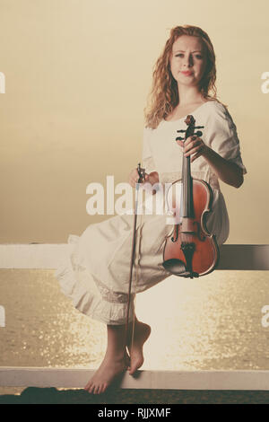 Musik liebe, Hobby und Alltag Leidenschaft Konzept. Frau auf Pier außerhalb Holding Violine bei Sonnenuntergang Stockfoto