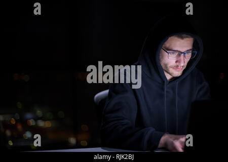 Junge talentierte Hacker mit Laptop Computers während des dunklen Büro arbeiten mit großen Lichter der Stadt im Hintergrund bei Nacht Stockfoto