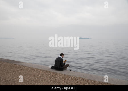 Ein junger Mann spielt ein Instrument, als er am 20. November 2018 an der Strandpromenade von Saloniki, Griechenland sitzt. Stockfoto