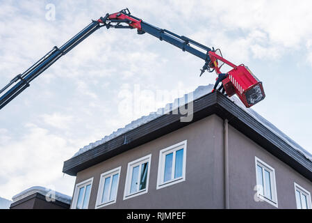Mann in die teleskopantenne Plattform Reinigung Schnee und Eis von Dach, dachrinne der Gebäude die Gefahr zu vermeiden. Stockfoto