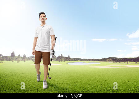 Stattliche asiatischer Mann in weißen Kleidern holding Eisen Golf Club zu Fuß auf den Golfplatz mit Bunkern, Teich und Bäume am Morgen Stockfoto