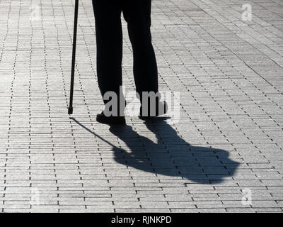 Silhouette der Mann mit einem Stock, lange Schatten auf Pflaster. Konzept der hinkt oder blind Person, Behinderung, Alter, Erkrankungen der Wirbelsäule Stockfoto