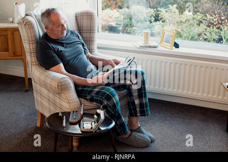 Ältere Diabetiker Mensch ist Entspannung im Wohnzimmer seines Hauses mit einem Buch. Sein Blut glucose Testing Kit ist auf den Tisch neben ihm. Stockfoto