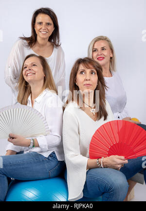 Gruppe von 4 Frauen, Freunden, im mittleren Alter Spaß haben in einer Fotosession in einem Studio mit weißem Hintergrund, sitzend auf einer Kugel und Spielen mit einem Ventilator Stockfoto