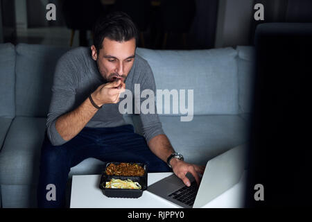 Junge Mann sitzt auf einem Sofa in Abend Essen zum Mitnehmen und mit Laptop Stockfoto