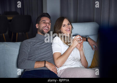 Junges Paar sitzt auf einem Sofa in Abend, Lachen während der TV-Programmwiedergabe Stockfoto