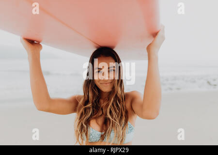 Junge weibliche Surfer, Surfbrett auf dem Kopf am Strand, Porträt, Ventura, Kalifornien, USA Stockfoto