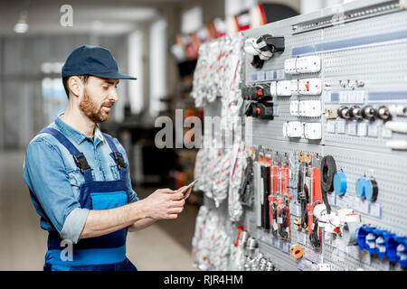 Stattliche Elektriker in der berufsbekleidung Auswahl elektrische Waren stehend mit Smart Phone im Gebäude shop Stockfoto