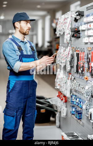 Stattliche Elektriker in der berufsbekleidung Auswahl elektrische Waren stehend mit Smart Phone im Gebäude shop Stockfoto