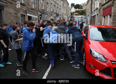 Die Jungs spielen beim jährlichen 'Fastern's E'en Hand Ba' Event auf der High Street in den Scottish Borders neben einem Auto um den Lederball. Stockfoto