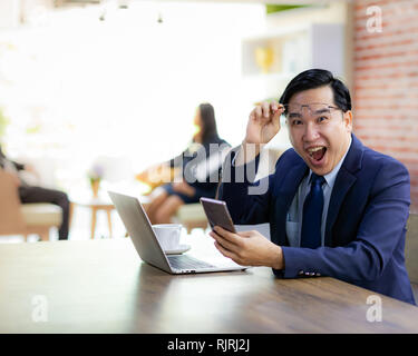 Portrait von Asiatische Geschäftsmann feiern und froh für seinen Erfolg im Cafe mit Smartphone und Laptop mit Business Team im Hintergrund für copor Stockfoto