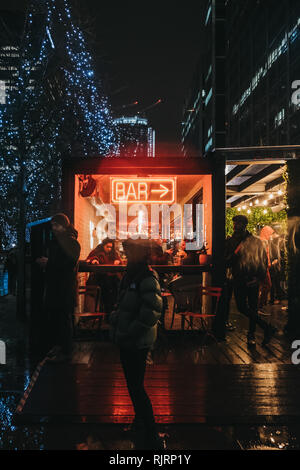 London, Großbritannien - 26 Januar, 2019: Menschen vorbei gehen. beleuchtete Fenster und Leuchtreklame auf Bar in Canary Wharf an einem regnerischen Abend. Canary Wharf ist ein Beschäftigter Stockfoto