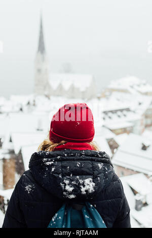 Frau in Red Hat genießt die Aussicht über Hallstatt Altstadt während der Schneesturm, Österreich. Ansicht von der Rückseite, vertikale Ausrichtung. Stockfoto
