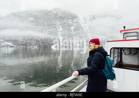 Frau in Red Hat steht auf einer Fähre Deck auf dem Weg nach Hallstatt, Österreich. Winter Blick auf den See und die Alpen. Stockfoto