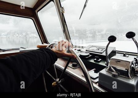 HALLSTATT, Österreich - Januar 2019: Blick über Hallstatt Stadt und Alpen von der Fähre Kapitänskajüte. Hand am Lenkrad. Stockfoto