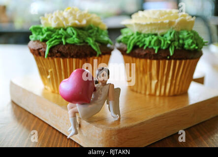 Geschlossen bis mini Engel holding Rosa großes Herz Figur mit ein paar Blumen geformte Schlagsahne schoko Cupcakes im Hintergrund Stockfoto