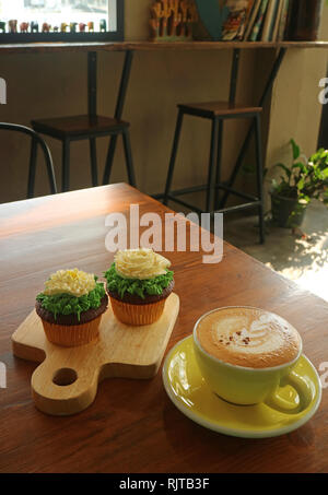 Tasse heißen Cappuccino und zwei Cupcakes gekrönt mit Blume geformte Schlagsahne in einem gemütlichen Raum serviert. Stockfoto