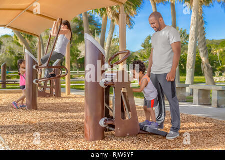 Ein Vater bringt seinen Sohn die Stairmaster, während Mutter und Tochter Fokus auf Pull-ups im Hintergrund verwenden. Die moderne Familie gemeinsam arbeiten Stockfoto