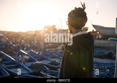 Jungen asiatischen Bärtigen touristische Mann mit Dreadlocks und Schal auf dem Kopf an der Blauen Fishermans Boote in Essaouira, Marokko am Abend Sonnenuntergang. Stockfoto