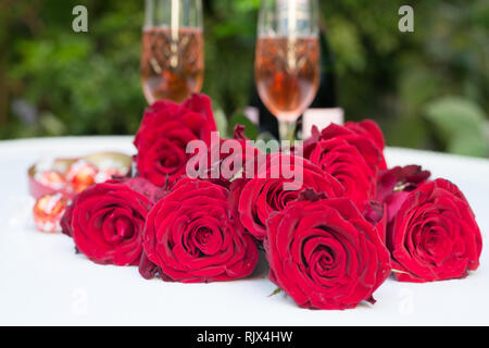 Rosen rot Bündel flach auf weißen Tisch im Freien mit zwei Gläser mit Champagner oder Sekt und Flasche plus container Pralinen gefüllt Stockfoto