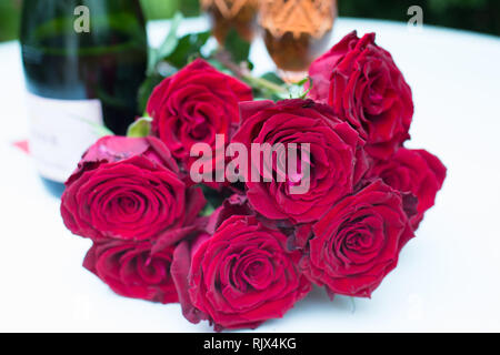 Rote Rosen strauss oder das Bouquet liegen auf weißen Tisch im Freien und im Hintergrund eine Flasche Champagner oder Sekt mit zwei Gläsern gefüllt Stockfoto