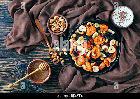 Getrocknete Früchte und Mutter Mix - Bananenscheiben, Aprikosen, Rosinen, Pflaumen, Kirschen und Cashew in einer Schüssel auf einem rustikalen Holztisch mit Honig in eine Schüssel geben und o Stockfoto