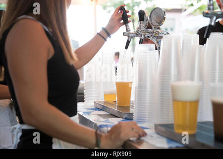 Nahaufnahme einer jungen Frau während Sie arbeiten am Zapfhahn. ist Sie tippen eine blonde Bier im Plastikbecher während ein Frühlingsfest. Stockfoto