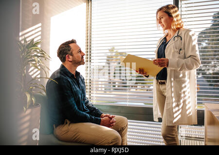 Ärztin, die ein ärztliches Gutachten Datei und im Gespräch mit männlicher Patient sitzt auf einem Sofa am Krankenhaus Rezeption. Arzt, der die Behandlung von Pa Stockfoto