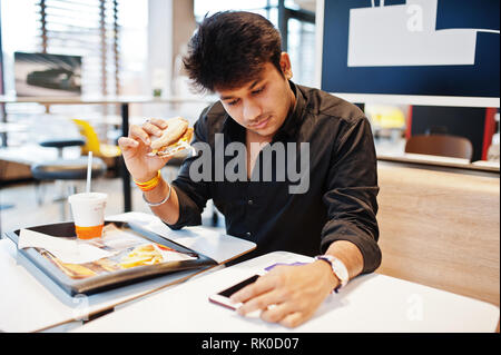 Stilvolle Inder in Fast-food Cafe und essen Hamburger sitzen, lesen Sie morgen News auf Handy. Stockfoto