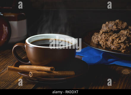 Dampfende Tasse Kaffee und Cinnamon Sticks mit einer Platte von Cookies im Hintergrund in einer dunklen Umgebung Stockfoto
