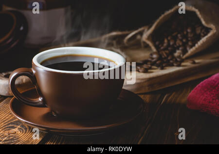 Nahaufnahme einer dampfenden Tasse Kaffee mit Kaffeebohnen verschüttete aus Sackleinen Tasche im Hintergrund in einer dunklen rustikalen Ambiente Stockfoto