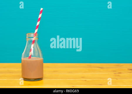 Ein Drittel pint Glas Milch Flasche halb voll mit Schokolade Milchshake mit retro Papier, Stroh, auf einen hölzernen Tisch gegen eine blaugrüne Hintergrund Stockfoto