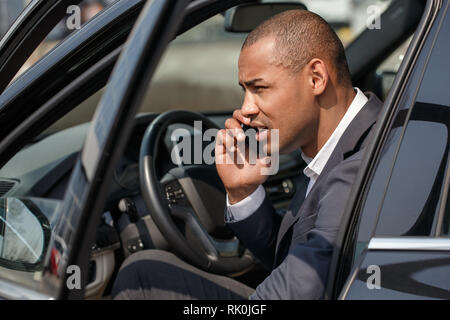 Junge afrikanische amerikanische Geschäftsmann sitzen im Auto Tür geöffnet in Konversation auf dem Smartphone beiseite suchen Schielen nachdenklich Stockfoto