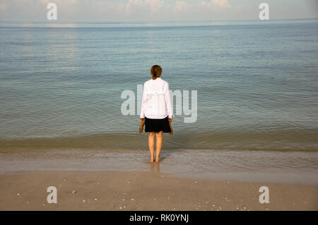 Halten Sie Ihre Sandalen, eine Frau steht in der Stille Ozean auf einem Strand in Naples, Florida, und leise blickt auf das Meer. Stockfoto