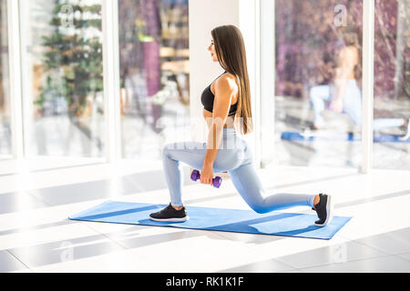 Tief in die Hocke. Seitenansicht der jungen schönen Frau in Sportkleidung tun Hocke und halten Hanteln beim Stehen vor der Fenster im Fitnessstudio. Stockfoto