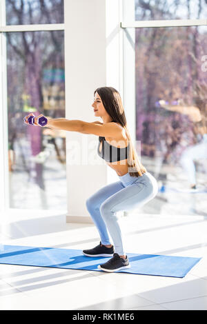 Tief in die Hocke. Seitenansicht der jungen schönen Frau in Sportkleidung tun Hocke und halten Hanteln beim Stehen vor der Fenster im Fitnessstudio. Stockfoto