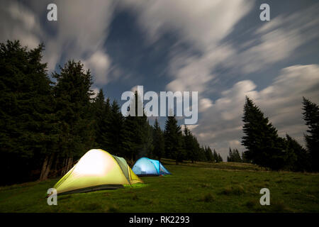 Zwei hell leuchtende, touristische Zelte auf der grünen Wiese Wald Lichtung unter hohen Kiefern auf klaren dunkelblauen Sternenhimmel Hintergrund. Tourismus, Nacht Camping Stockfoto