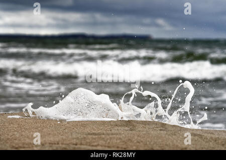 Eine kleine Welle bricht auf der Ostsee Strand vor einem kontrastierenden, Himmel, Spray und einem Tropfen Wasser vom Untergrund abheben - Loca Stockfoto