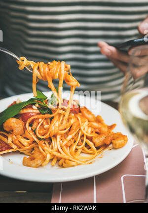 Italienisches Abendessen im Bistrot mit spaghetti Pasta mit Garnelen und Glas Weisswein. Mann essen Pasta und Smartphone. Köstliche italienische cui Stockfoto