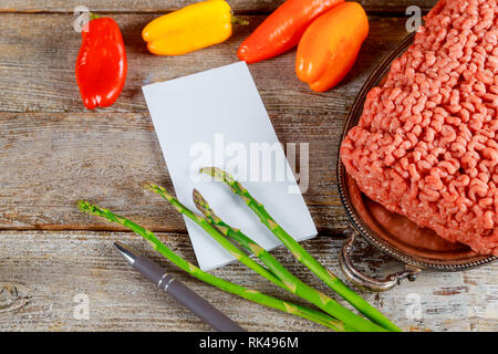 Rindfleisch Hackfleisch mit Gewürzen und Gemüse, bunte Paprika und Spargel, Notebooks, Pen auf alten Holz Hintergrund. Stockfoto