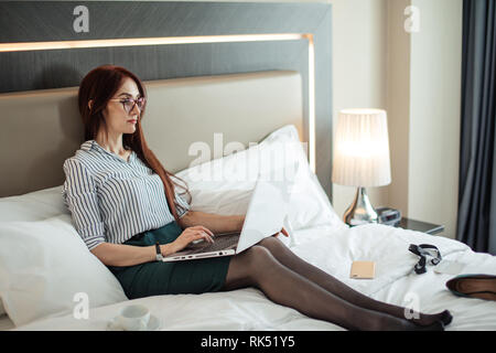 Erfolgreiche weibliche Rechtsanwalt ruht in diesem Hotel während einer Geschäftsreise. Rothaarige Frau liegt auf breiten Bett mit einem Laptop, elegant gekleidet Stockfoto