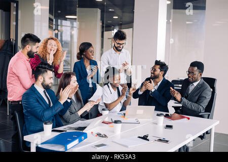 Mixed-Race Gruppe von neun verschiedenen glückliche Menschen stehen rund um die Tabelle mit den Kollegen sitzen, applaudieren Team Leistungen Stockfoto