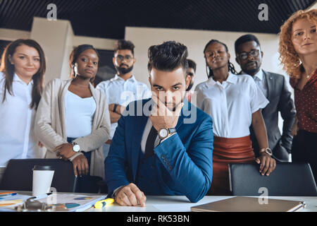 Creative smart Leader mit erweiterten Ziele. Aussehenden bärtigen Boss berühren sein Kinn, während seine Kollegen hinter ihm Stockfoto