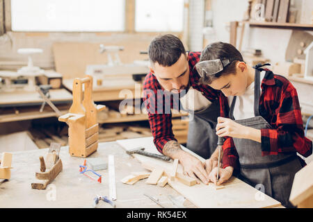 Bärtige Vorschäler erwachsenen Mann beobachtete, wie seine Jungen kleinen Sohn in eine Schutzbrille und eine Schürze arbeitet mit Meißel in eine Schreinerei. Stockfoto