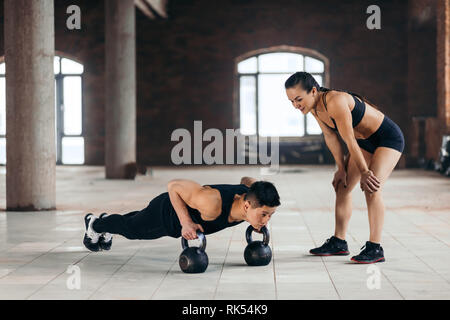 Weibliche personal trainer Controlling ein Mann, wer Push-ups mit den Kettlebells. Frau ermutigt ihren Freund im für den Sport zu gehen Stockfoto