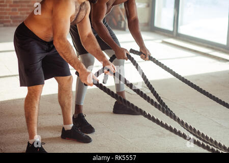 Zwei multiethnischen atletic Freunde der Stärke von einem unsichtbaren Feind mit Schlacht Seil in crossfit style Fitness Test, stehend mit Beine auseinander, Schulter - wid Stockfoto