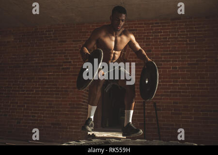 African American crossfit Sportler Holding barbell Platten als Gewichtung in beiden Händen und springt auf Große Gummiräder, liegen auf dem Boden der Straße l Stockfoto