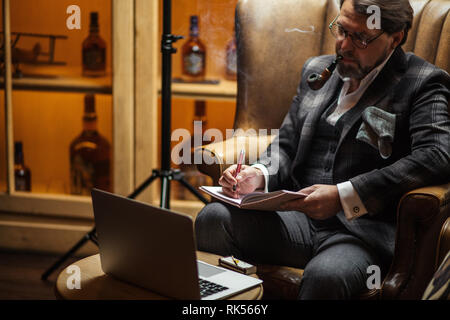 Gut aussehender Mann mittleren Alters blogger tragen trendy Tweed Jacke, puffing Tabakpfeife und Arbeiten an Cafe Tabelle mit Laptop, Schreiben in neuer Artikel Stockfoto