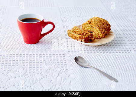 Eine rote Tasse mit Kaffee auf eine weiße Tischdecke, ein süßes Brötchen auf dem Teller Stockfoto