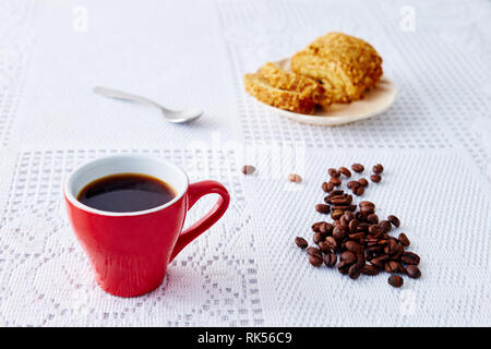 Eine rote Tasse mit Kaffee auf eine weiße Tischdecke, ein süßes Brötchen auf dem Teller Stockfoto